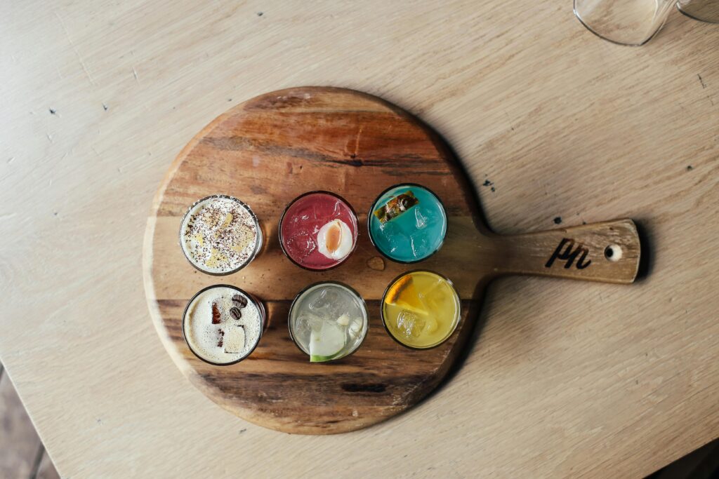 Top view of six colorful cocktails arranged on a wooden serving board, perfect for a bar menu or beverage display.